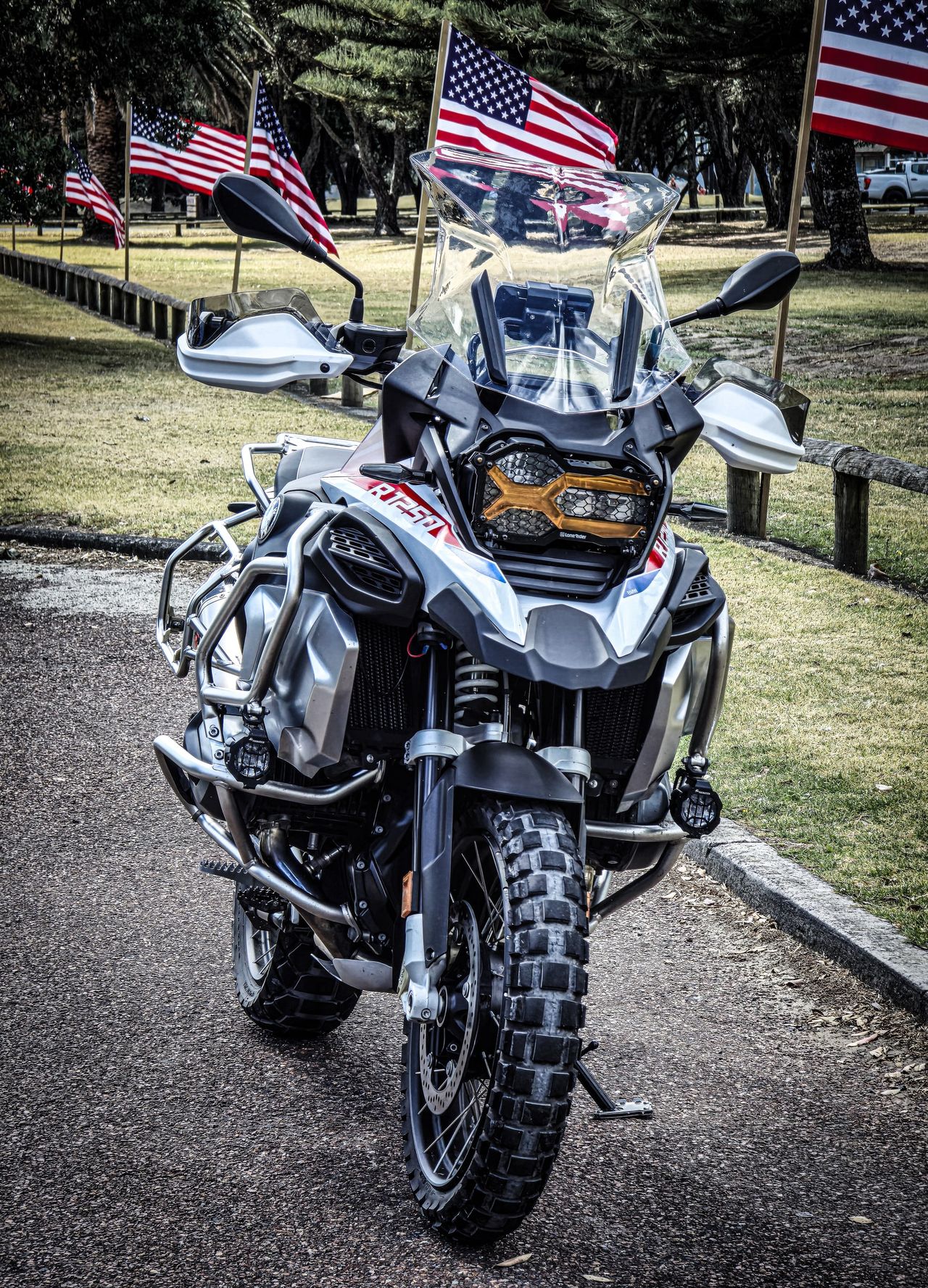 Front view of a white, blue and red BMW R 1250 GS Adventure with knobbly tyres and an amber headlight guard, US flags behind