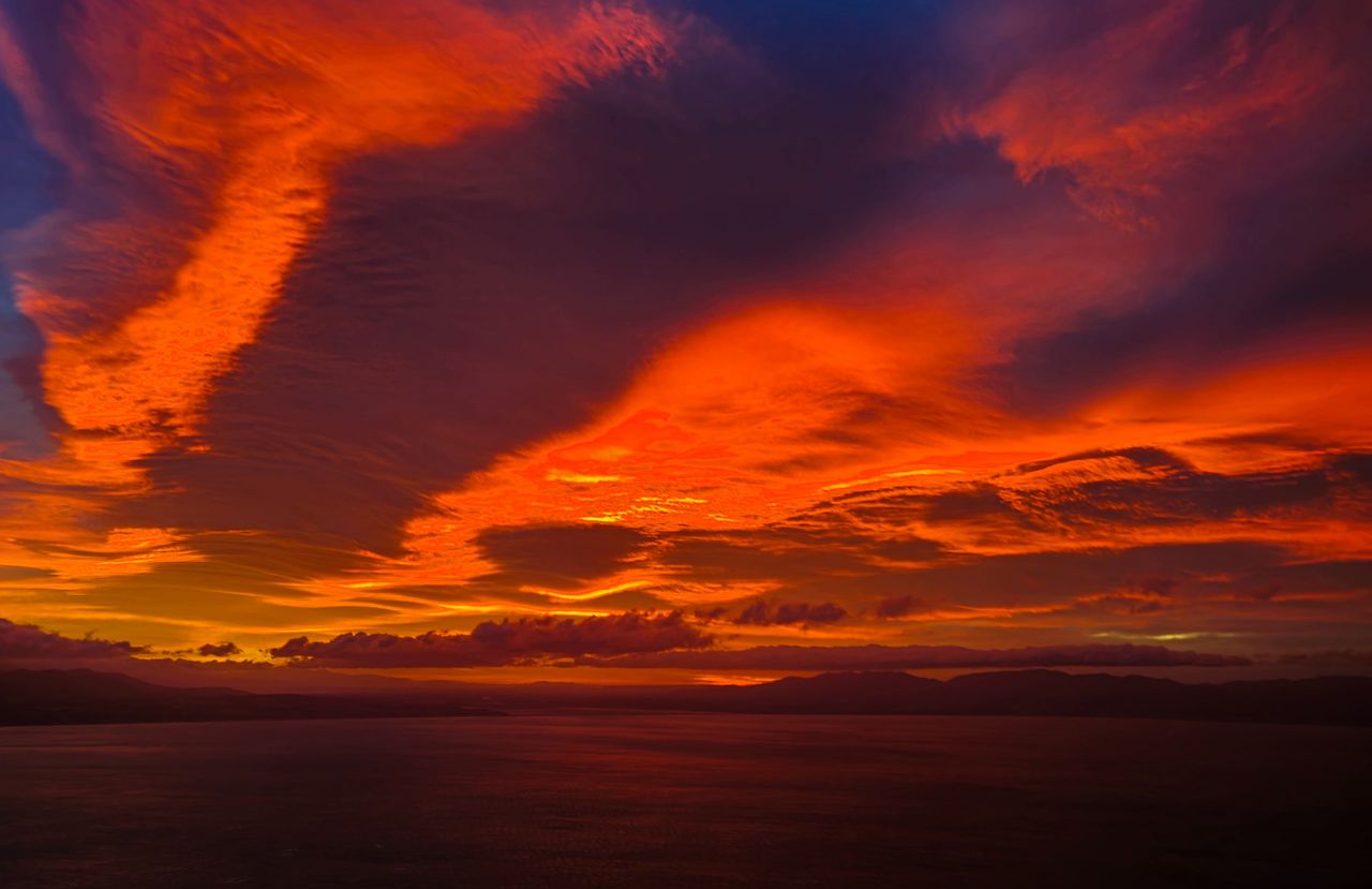 Vivid red and orange clouds streaking across a dawn sky above dark, calm water and a low line of hills