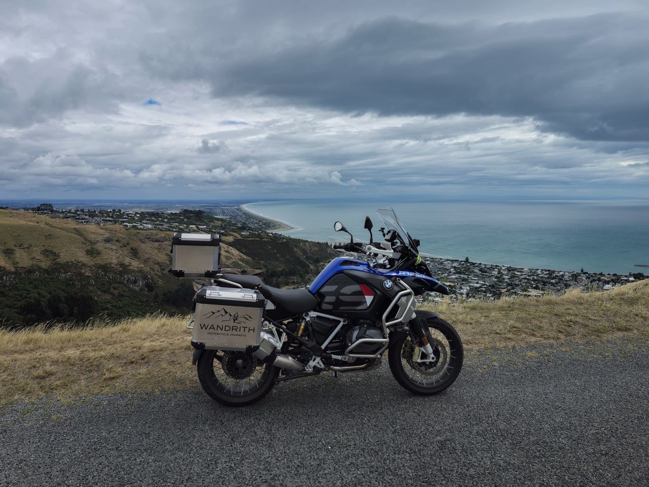 Blue BMW R 1250 GS Adventure with Wandrith panniers on a hilltop road above a seaside suburb, a long curving beach and grey cloud