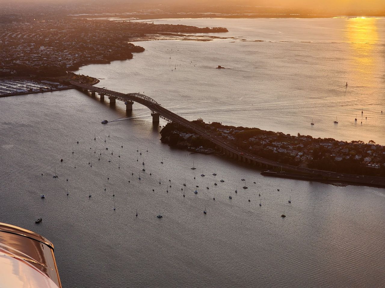 Aerial view at sunset of the Auckland Harbour Bridge over the Waitematā, moored yachts below, part of the aircraft in the corner