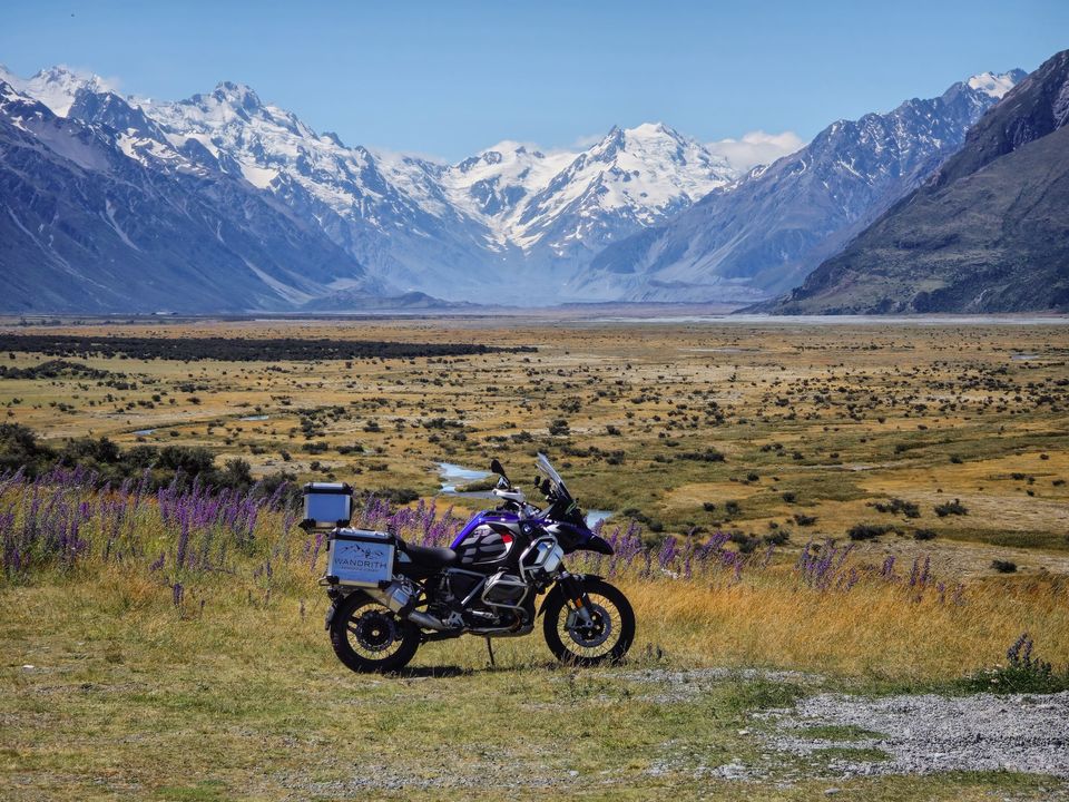 BMW adventure motorcycle with Wandrith panniers among purple lupins and golden tussock, a wide braided valley and snowy peaks beyond