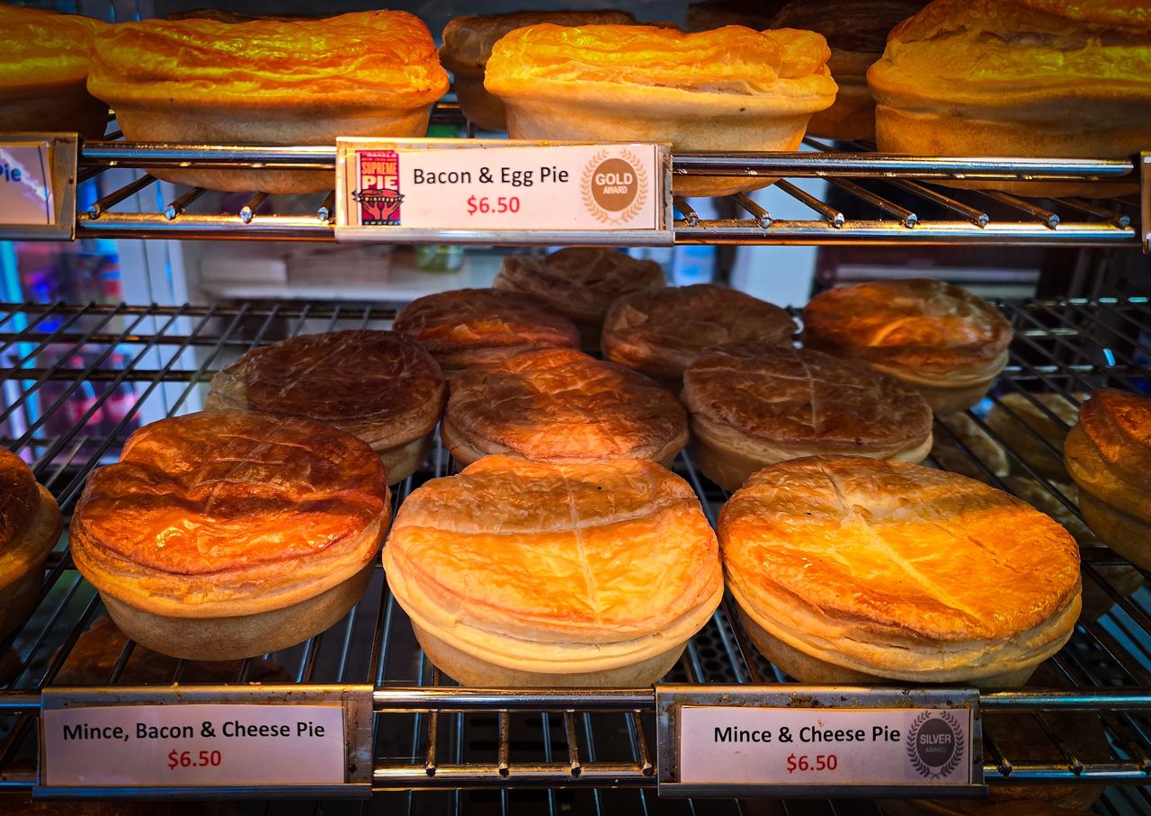 Rows of golden, flaky pies on wire shelves in a bakery warmer, labelled bacon and egg, mince and cheese, and mince, bacon and cheese
