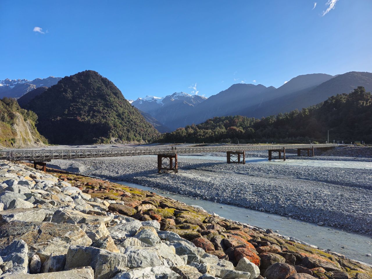 Wide grey gravel riverbed crossed by a long narrow bridge, with bush-covered hills and snowy peaks under a clear blue sky