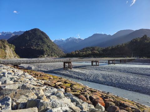Wide grey gravel riverbed crossed by a long narrow bridge, with bush-covered hills and snowy peaks under a clear blue sky