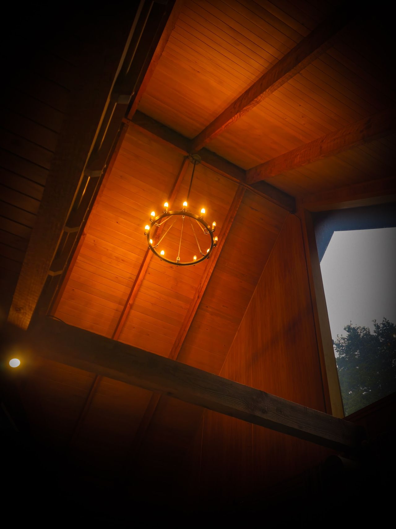Looking up at a vaulted timber ceiling with exposed beams and a ring chandelier of candle-style bulbs, a tall window onto trees