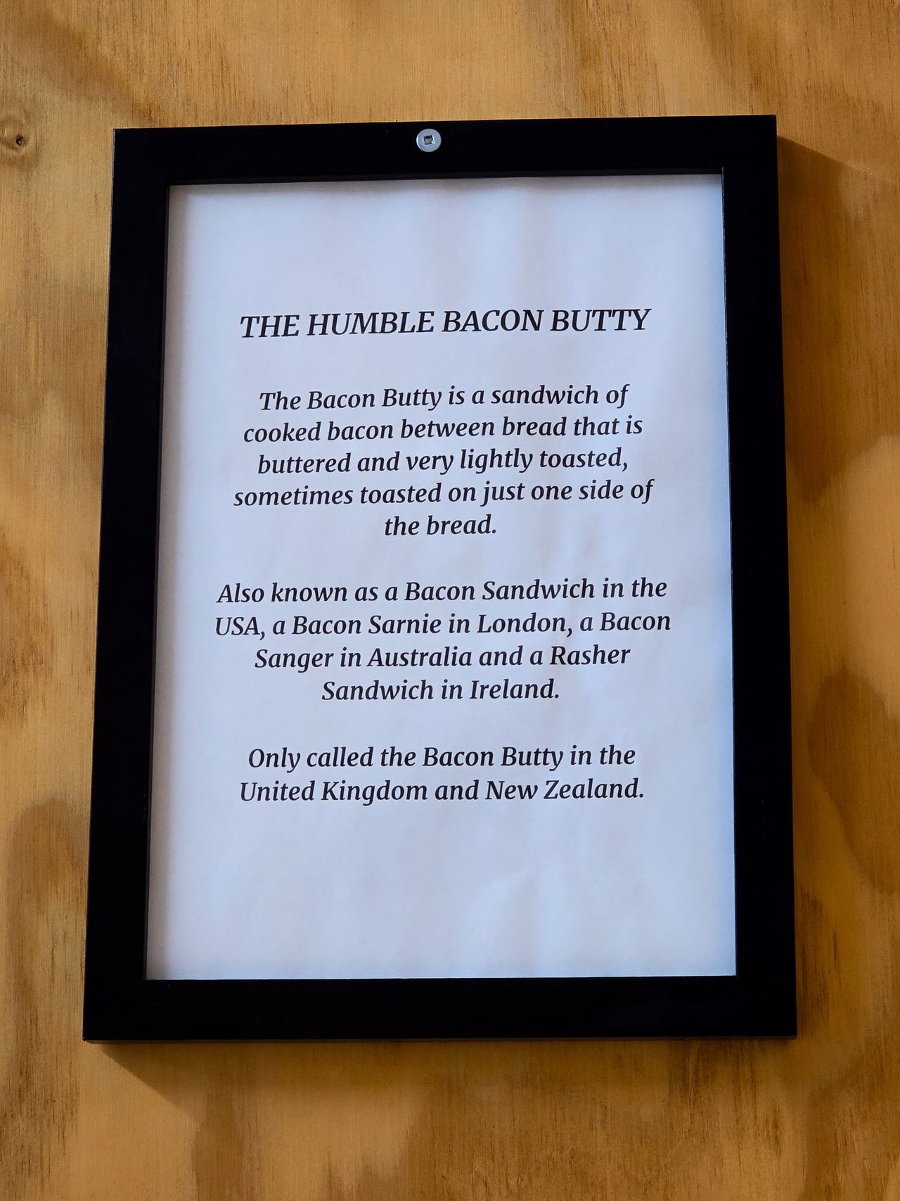 Framed sign on a plywood wall headed 'The Humble Bacon Butty', explaining the sandwich and its names in other countries