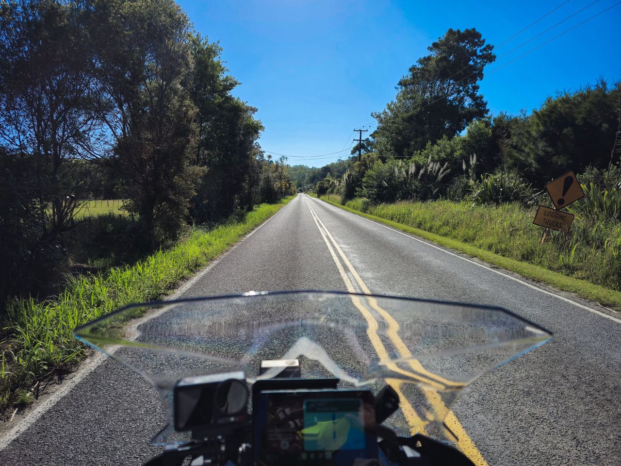 Rider's-eye view over a motorcycle windscreen along a straight country road lined with bush and flax, past a logging trucks warning sign