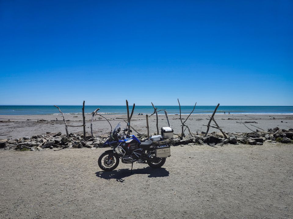 BMW R 1250 GS Adventure with Wandrith panniers by driftwood poles on a stony beach, open sea under a cloudless sky