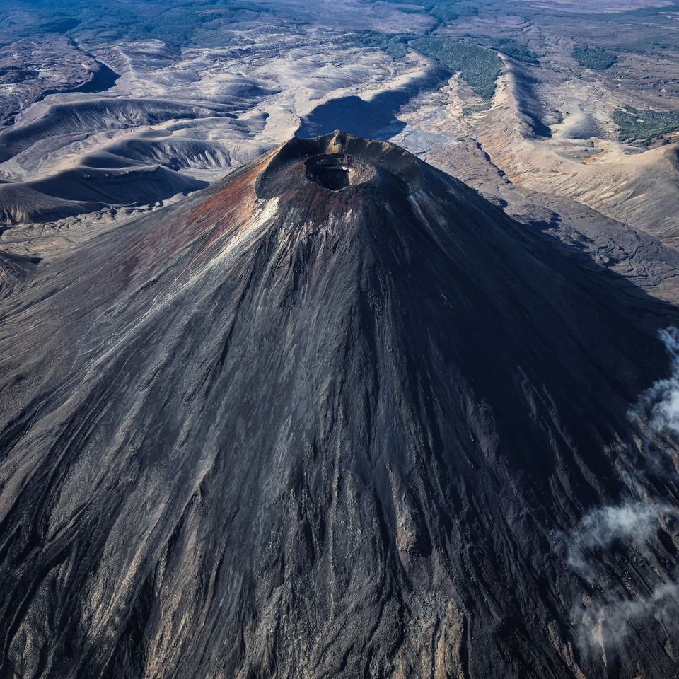 Aerial view down onto the steep, dark cone of a volcano with a summit crater, surrounded by barren lava fields
