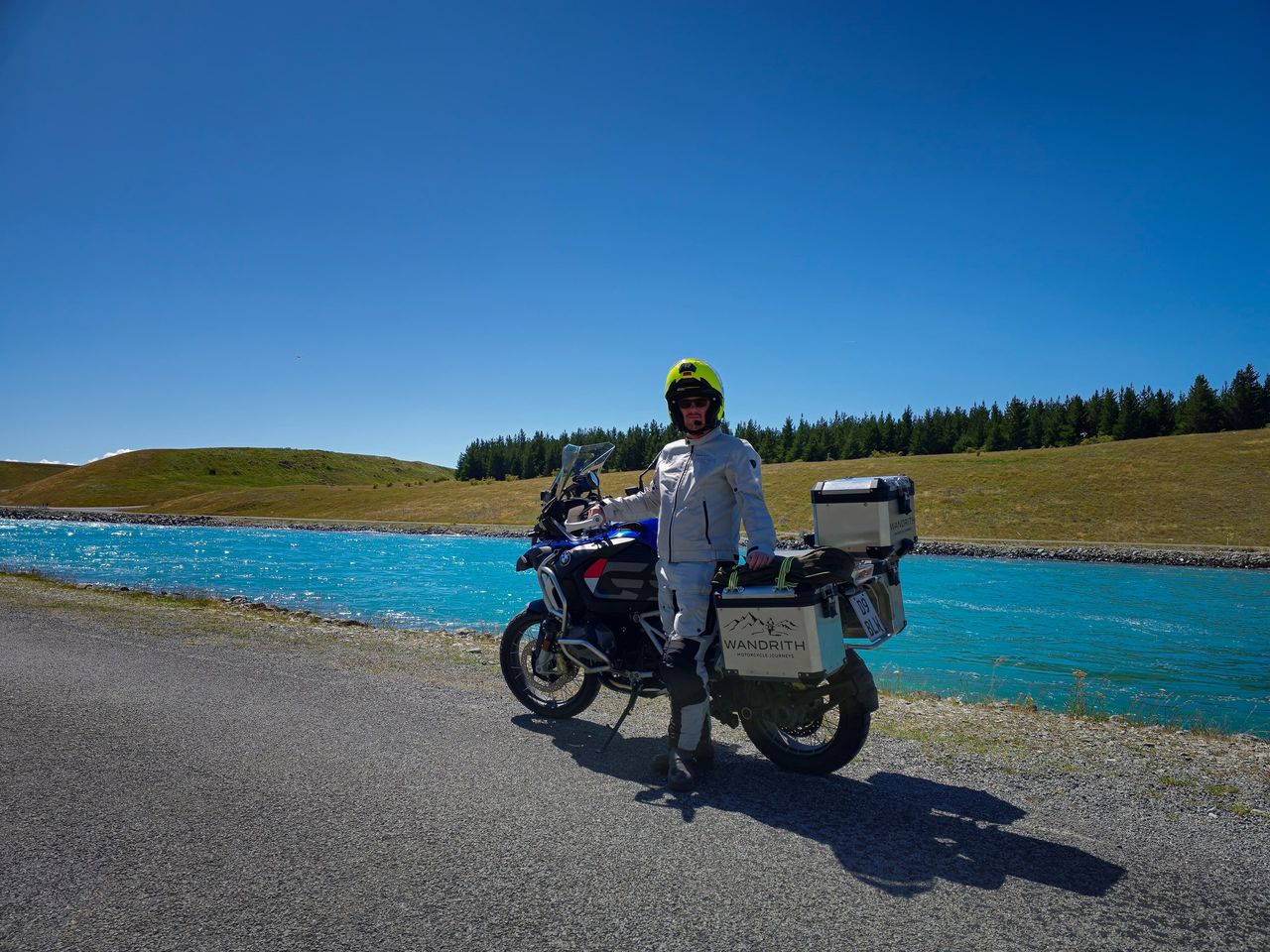 Rider in a light grey suit and yellow helmet beside a loaded BMW adventure motorcycle with Wandrith panniers, next to a turquoise canal