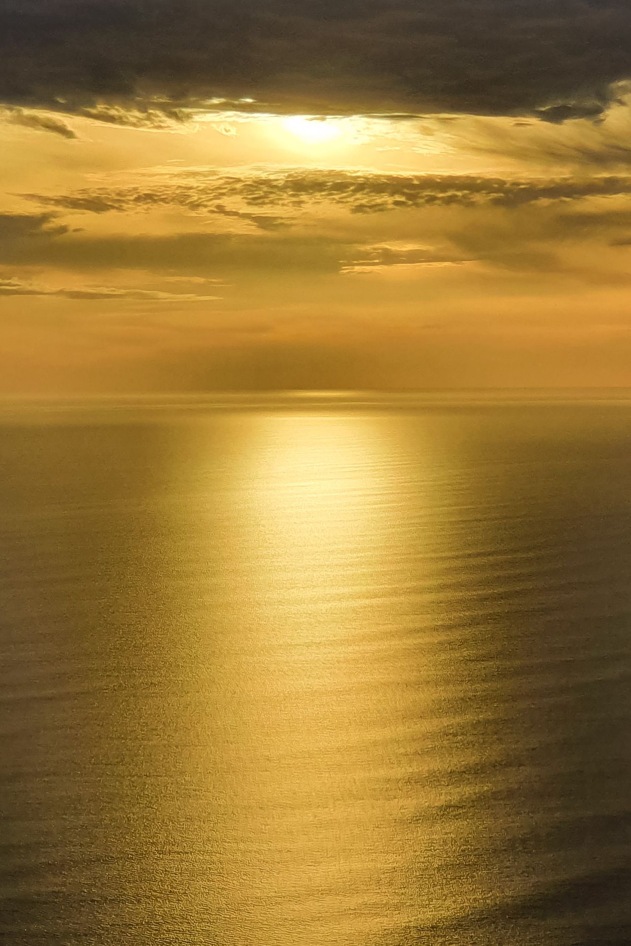 Late-afternoon sun breaking through cloud above a calm sea, casting a long golden path across the water