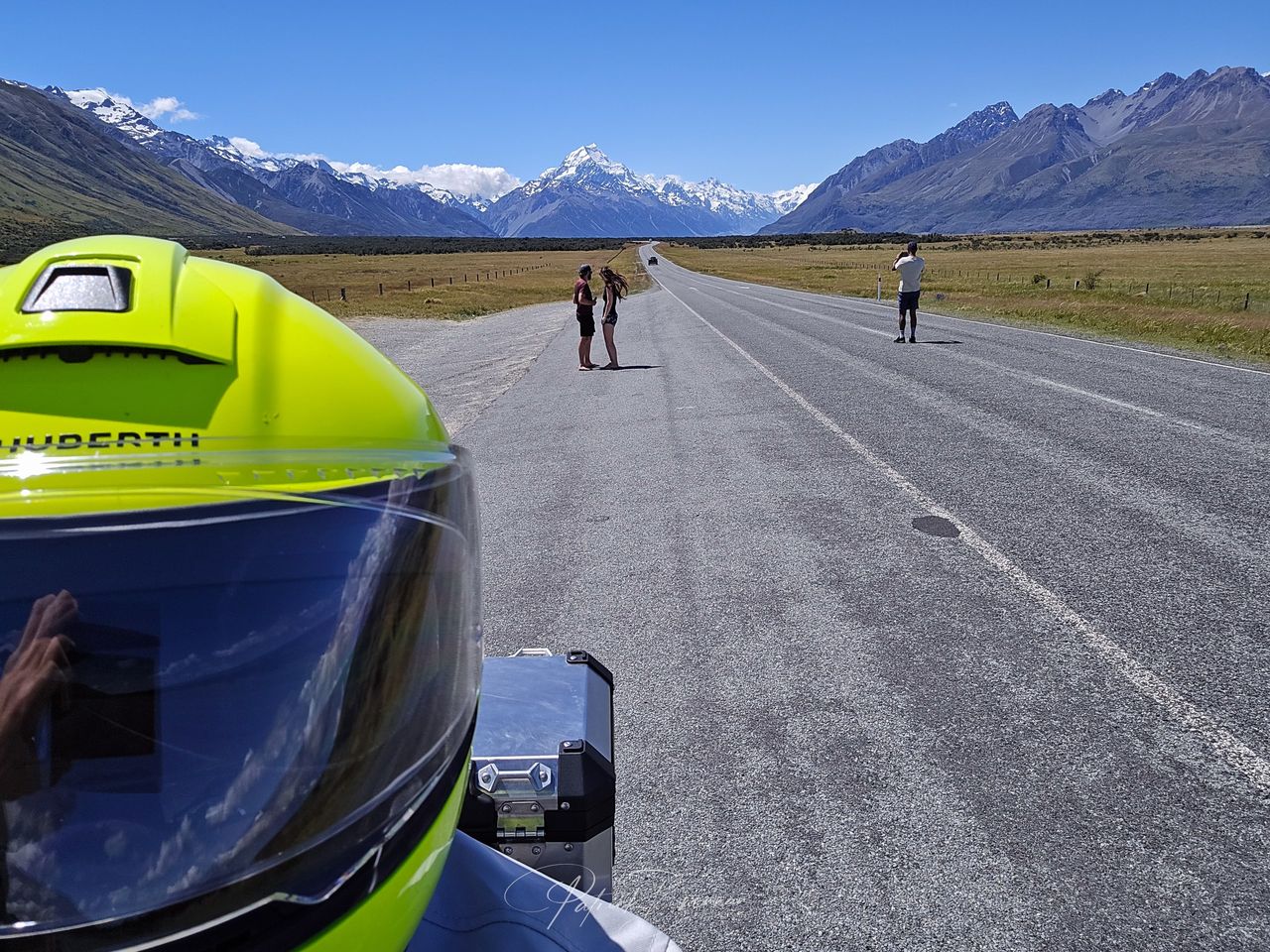 Rider’s view past a hi-vis helmet down a long straight road to snowy Aoraki / Mount Cook, with people stopped for photos