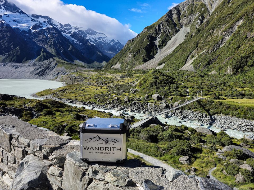 Wandrith top box on a stone wall above a milky glacial lake, a rocky river and a swing bridge, with snowy peaks behind