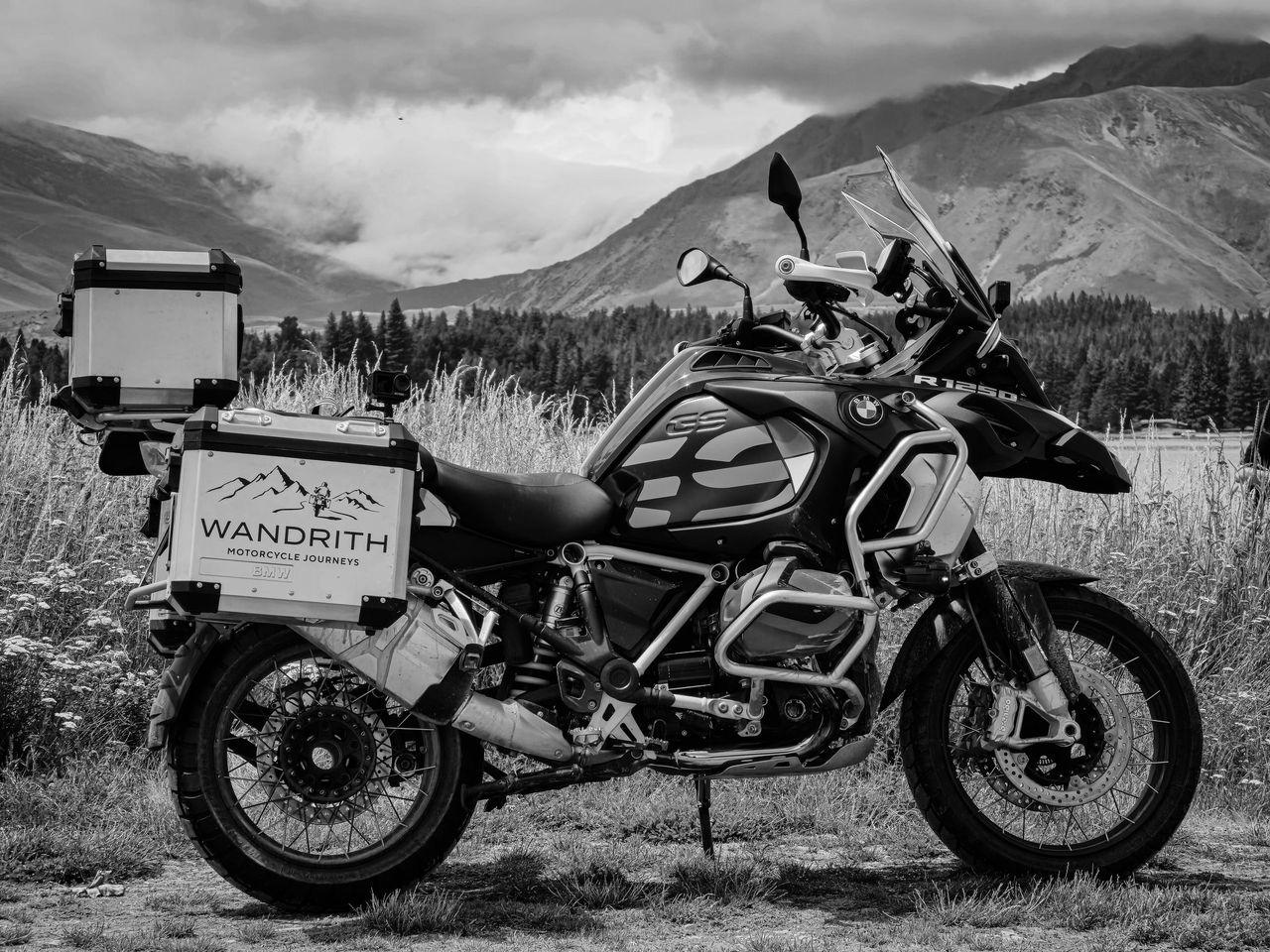Black-and-white photo of a BMW R 1250 GS Adventure with Wandrith panniers and top box in long grass, forest and cloudy mountains behind