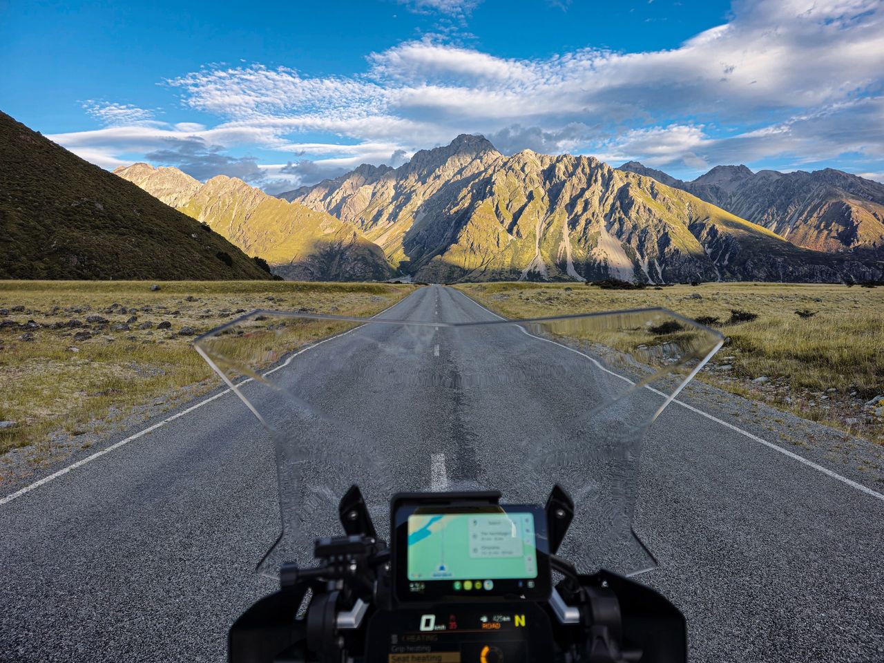 Rider's-eye view over a motorcycle windscreen and map screen, down an empty road through tussock toward sunlit, rugged mountains