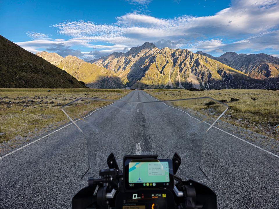 Rider's-eye view over a motorcycle windscreen and map screen, down an empty road through tussock toward sunlit, rugged mountains