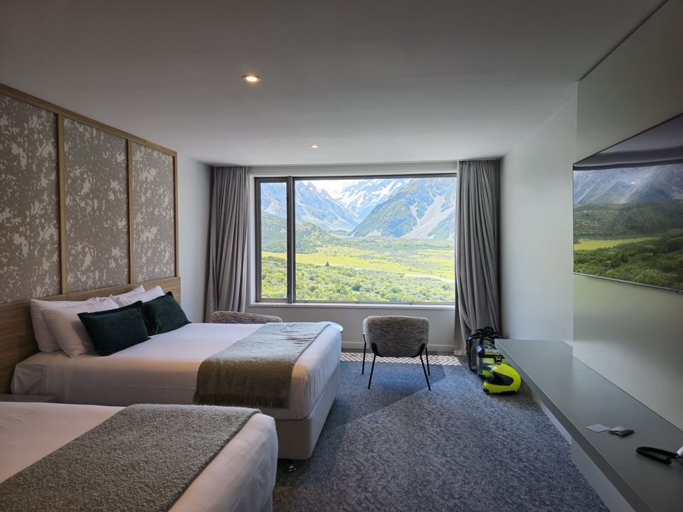 Hotel room with two beds and a picture window framing a green glacial valley and snowy peaks; a rider’s helmet on the floor