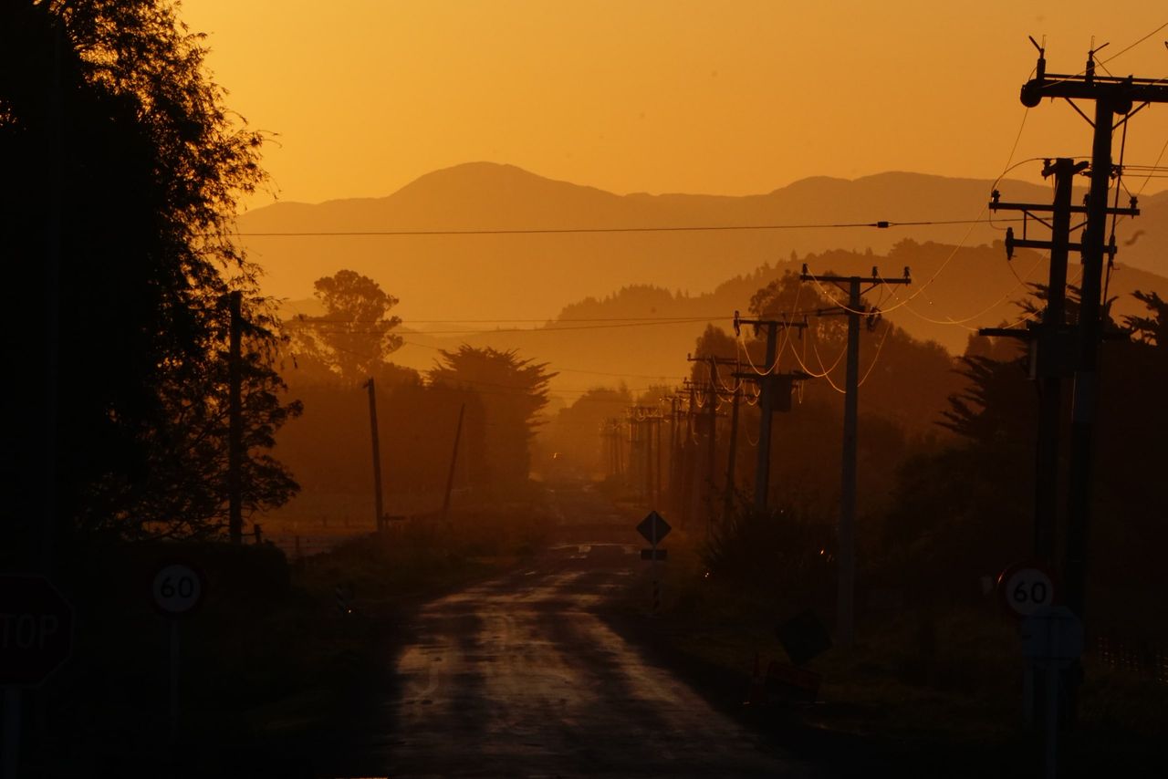 Narrow country road at sunset running toward hazy hills, lined with power poles, trees and 60 km/h signs, all glowing orange