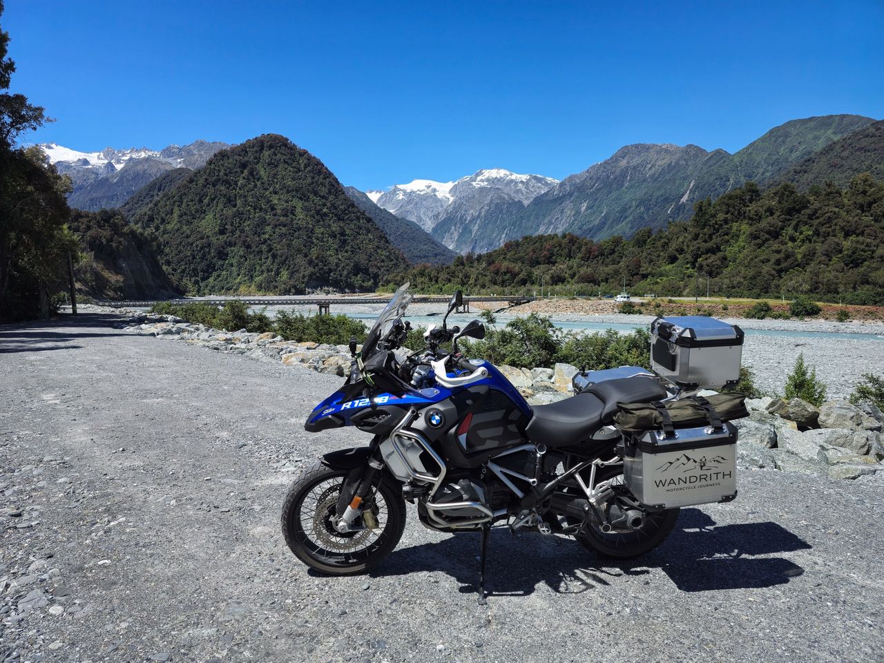 Blue BMW R 1250 GS Adventure with Wandrith panniers by a wide stony riverbed and single-lane bridge, rainforest hills and snowy peaks behind