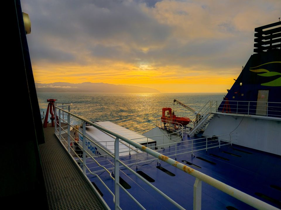Upper deck of a ferry at sunrise: railings, an orange rescue boat and a truck trailer, with golden light over the sea and distant hills