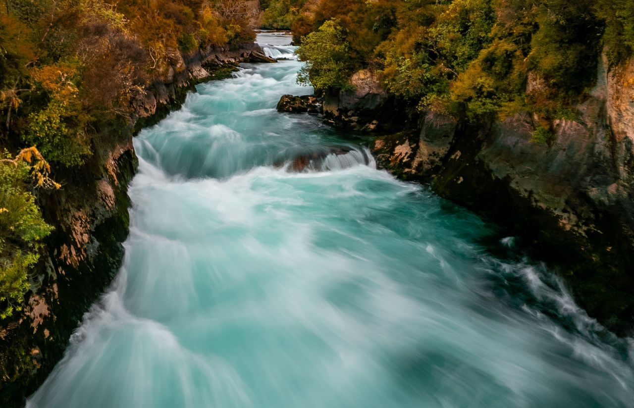 Long-exposure view of turquoise water surging white through a narrow rock channel lined with autumn-tinted bush