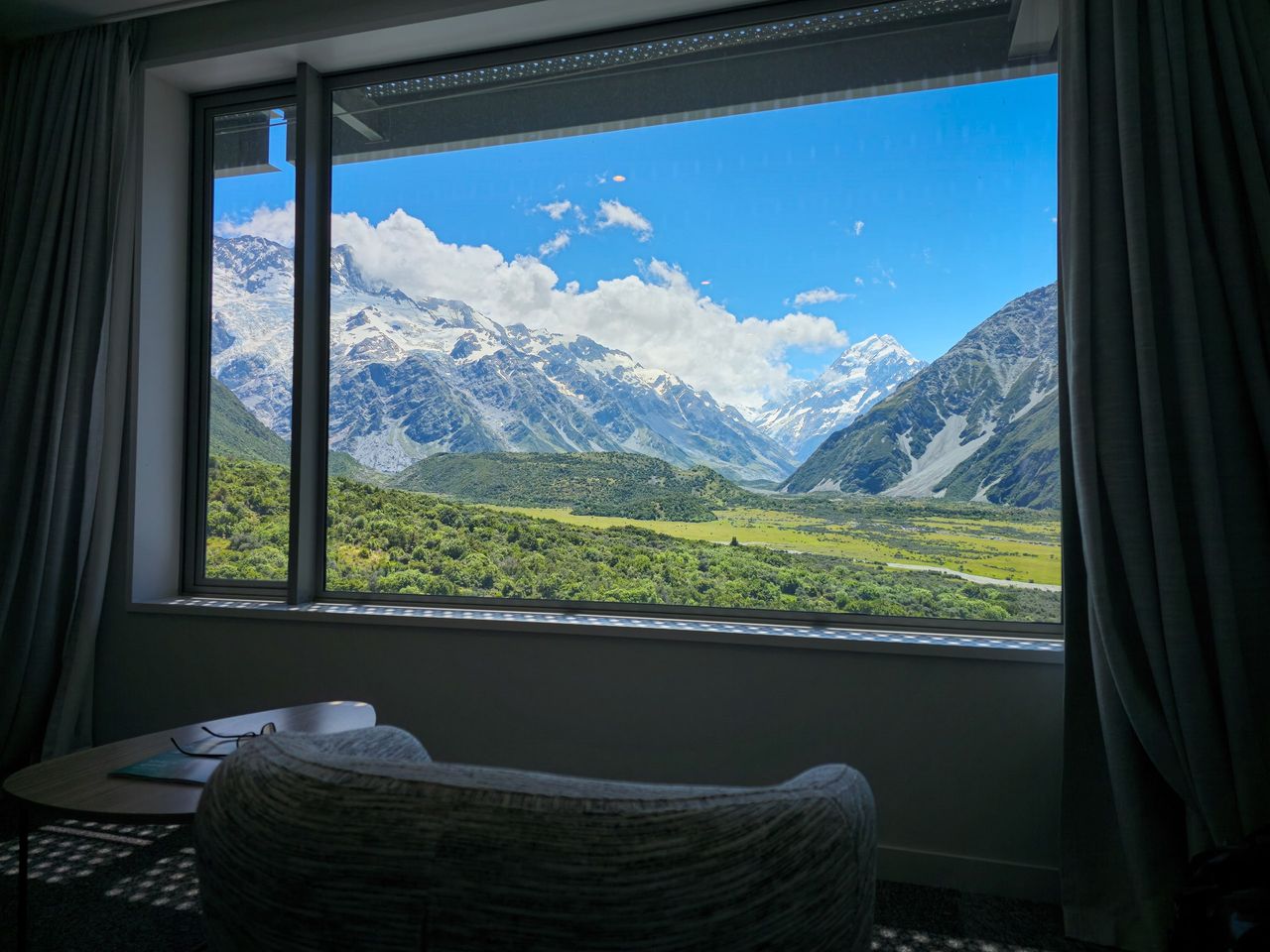 Hotel room window framing Aoraki / Mount Cook and snowy peaks above a green valley floor, an armchair and small table in front