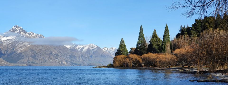Blue lake with snow-capped mountains on the left and a shoreline of tall conifers and leafless golden willows on the right