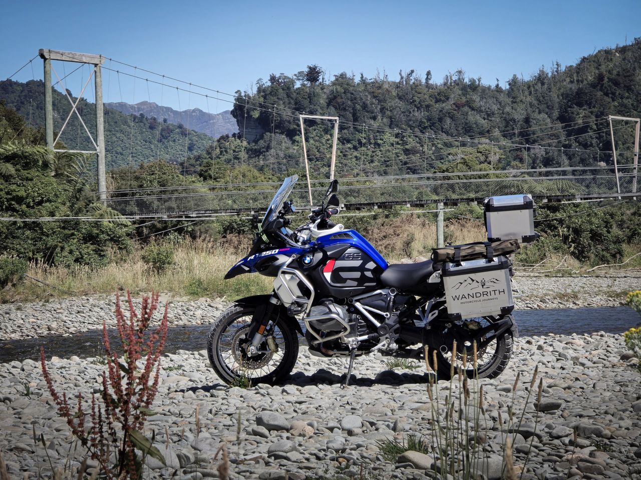 BMW R1250GS Adventure with Wandrith panniers on a stony riverbed beside a suspension footbridge, native bush and rocky bluffs behind