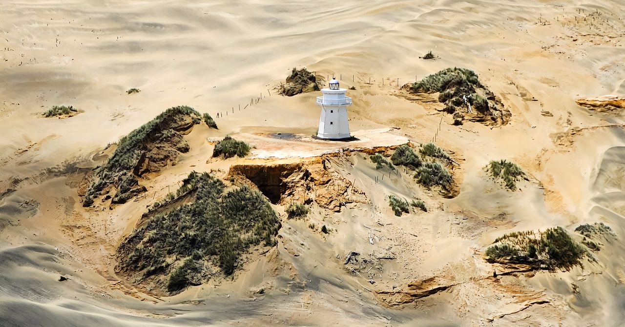 Small white lighthouse on an eroded sandy rise, ringed by wind-rippled dunes and grassy hummocks, seen from above