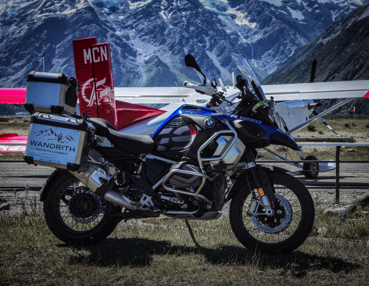 BMW R 1250 GS Adventure with Wandrith panniers parked beside a red-and-white light aircraft, glaciated mountains behind