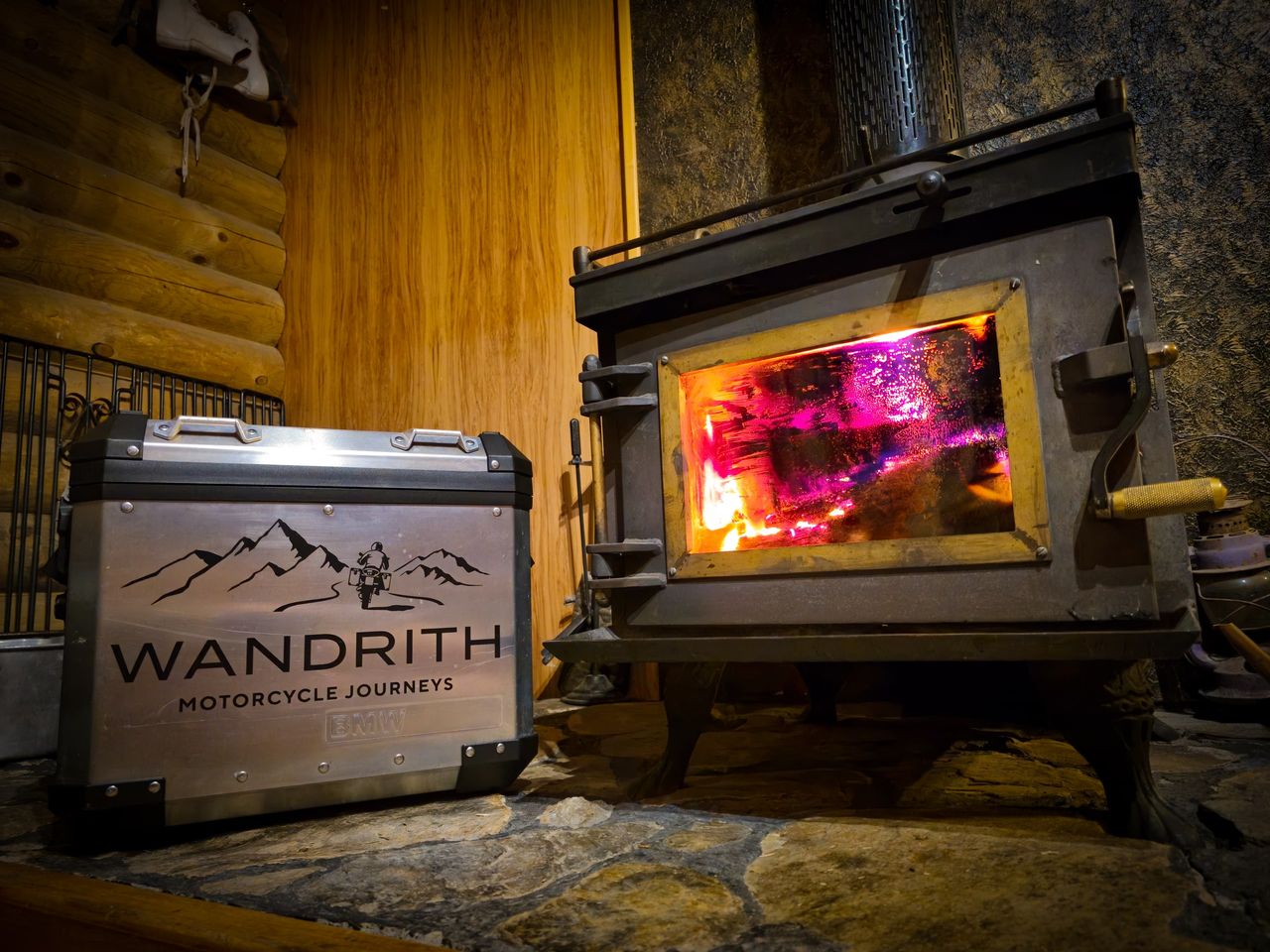 Aluminium BMW pannier with the Wandrith Motorcycle Journeys logo on a stone hearth beside a cast-iron wood burner, log walls behind