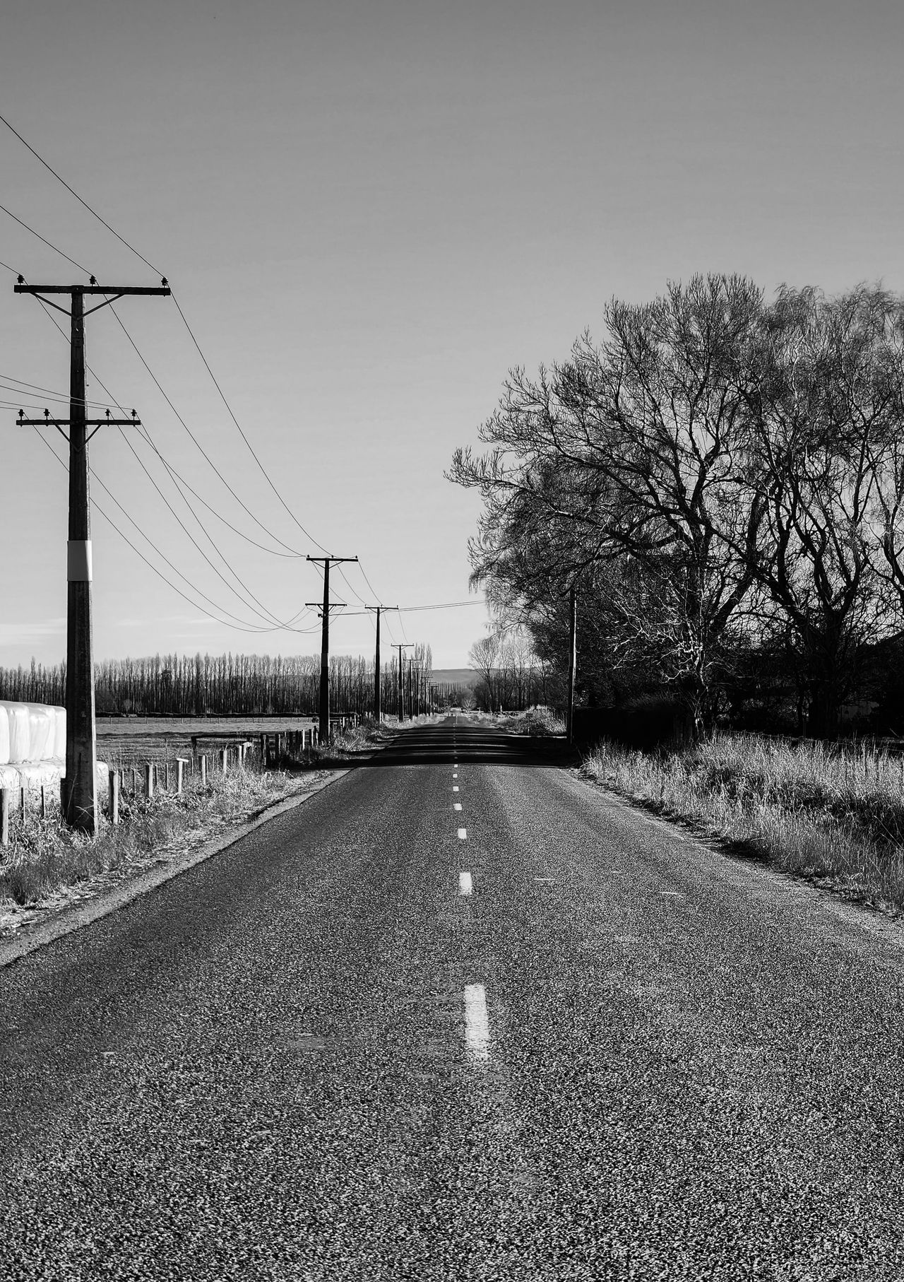 Black-and-white photo of a straight country road running into the distance past power poles, wrapped bales and bare winter trees