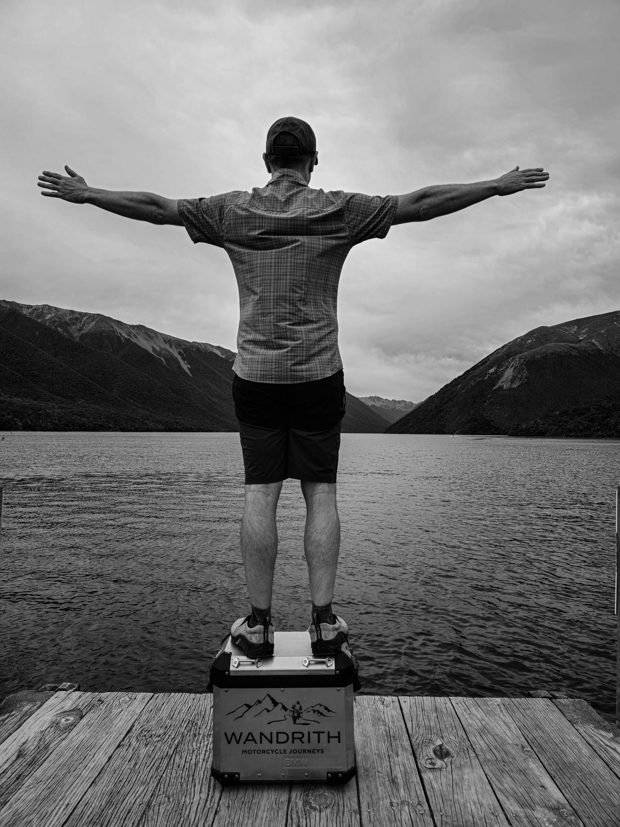 Black-and-white: a person seen from behind, arms outstretched, standing on a Wandrith pannier on a jetty, facing a lake and steep mountains