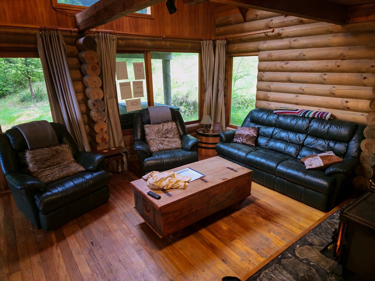 Log cabin lounge with dark leather recliners and sofa, a wooden chest as coffee table, a wood burner and windows onto bush