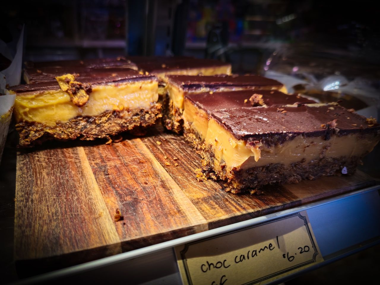 Thick squares of chocolate caramel slice on a wooden board in a café cabinet, with a handwritten price label in front