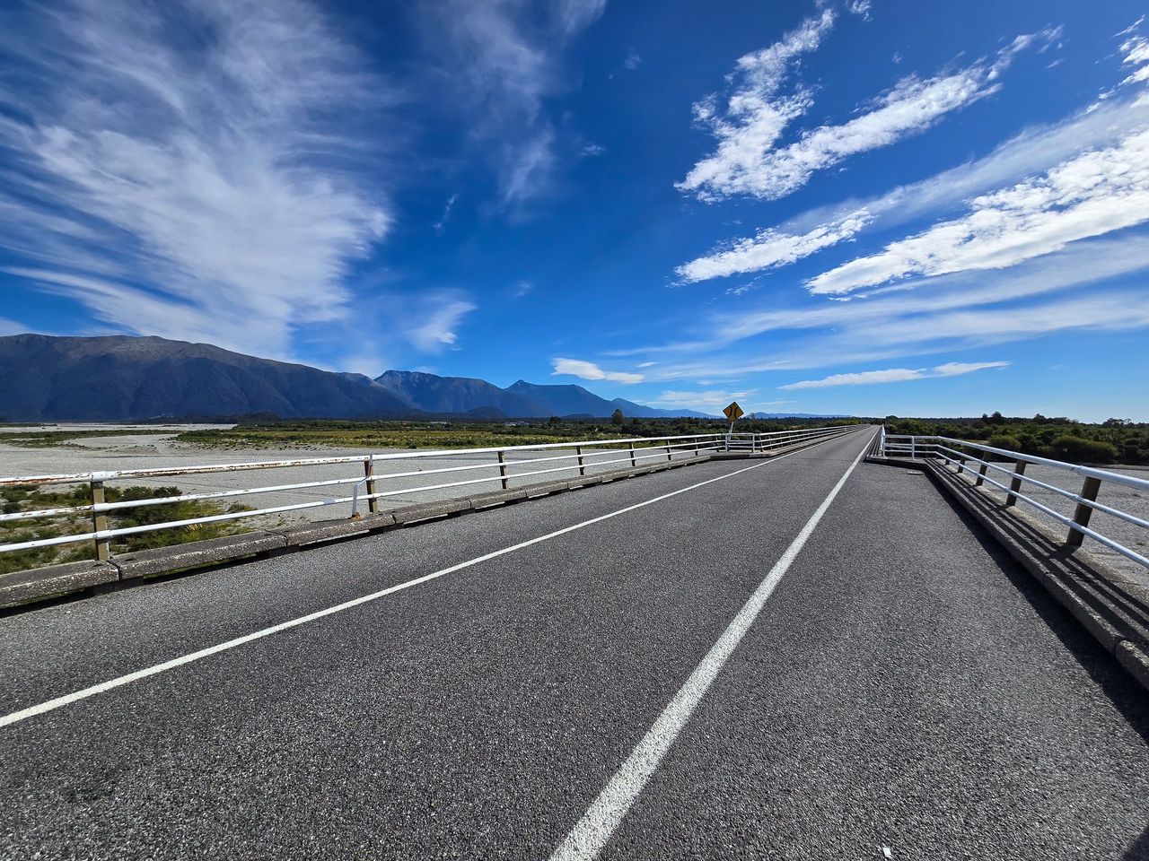 Low wide-angle view along a long road bridge over a broad braided riverbed, with bush-covered ranges and streaky cloud