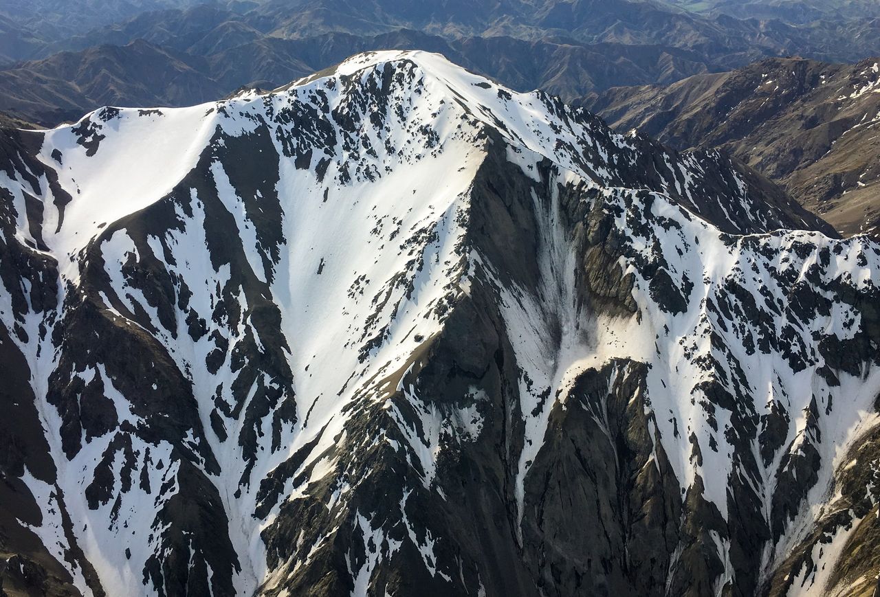 Aerial view of a steep mountain ridge streaked with snow and dark rock, with brown, mostly snow-free ranges behind