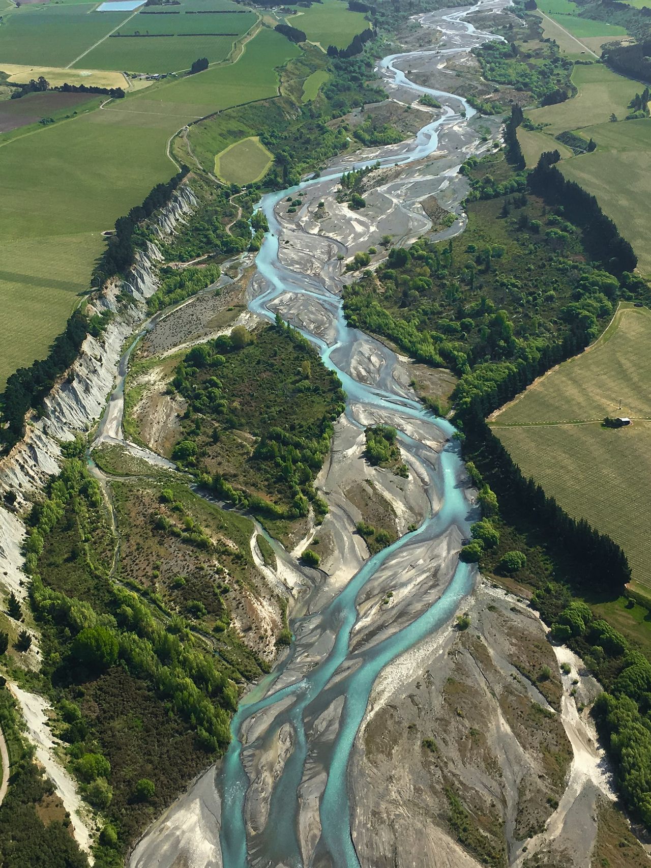 Aerial view of a turquoise braided river winding through grey gravel beds, with pale cliffs and green farmland on both sides