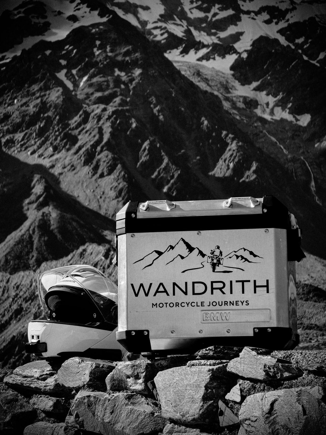 Black-and-white shot of a Wandrith aluminium top case and white helmet on rocks, a craggy glaciated mountainside behind