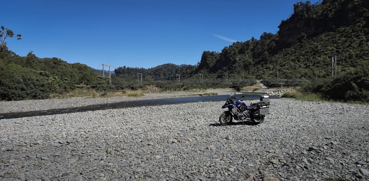 Loaded adventure motorcycle parked on a wide stony riverbed, a long swing bridge and bush-covered cliffs behind, blue sky