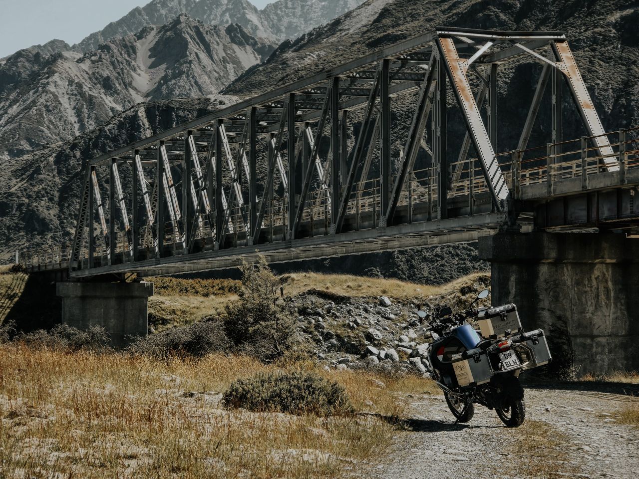 Loaded BMW GS Adventure on a gravel track beside an old steel truss bridge, with rocky mountains and tussock behind