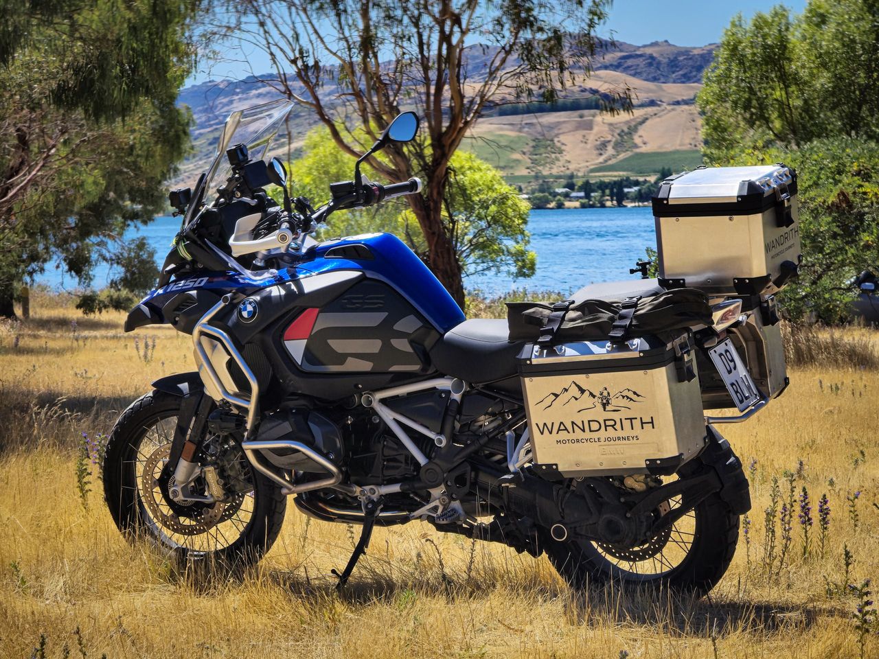 Blue and black BMW R 1250 GS Adventure with Wandrith luggage on golden grass under trees, a blue lake and dry hills behind