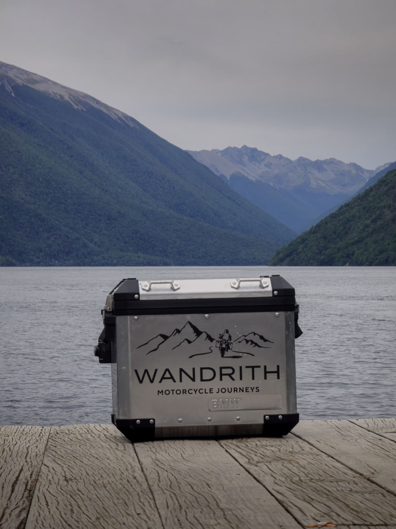 Silver BMW pannier with the Wandrith Motorcycle Journeys logo on a weathered jetty, a lake and forested mountains behind