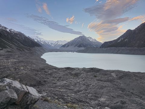 Pale glacial lake between grey moraine walls at dusk, snow-capped peaks at the head of the valley and pink-lit clouds