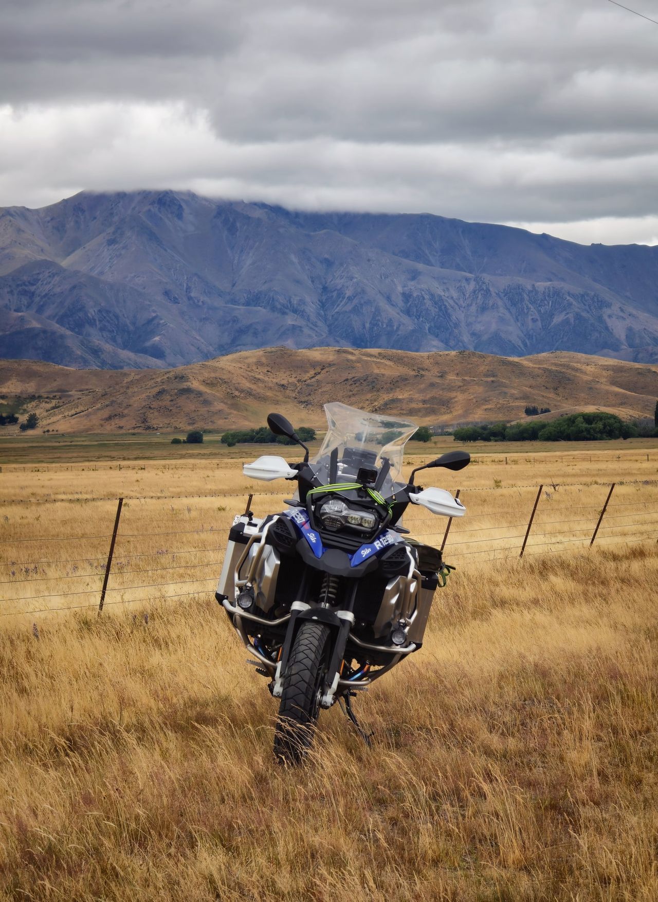 BMW adventure motorcycle with panniers, head-on in long golden grass by a wire fence, brown hills and cloud-capped mountains behind