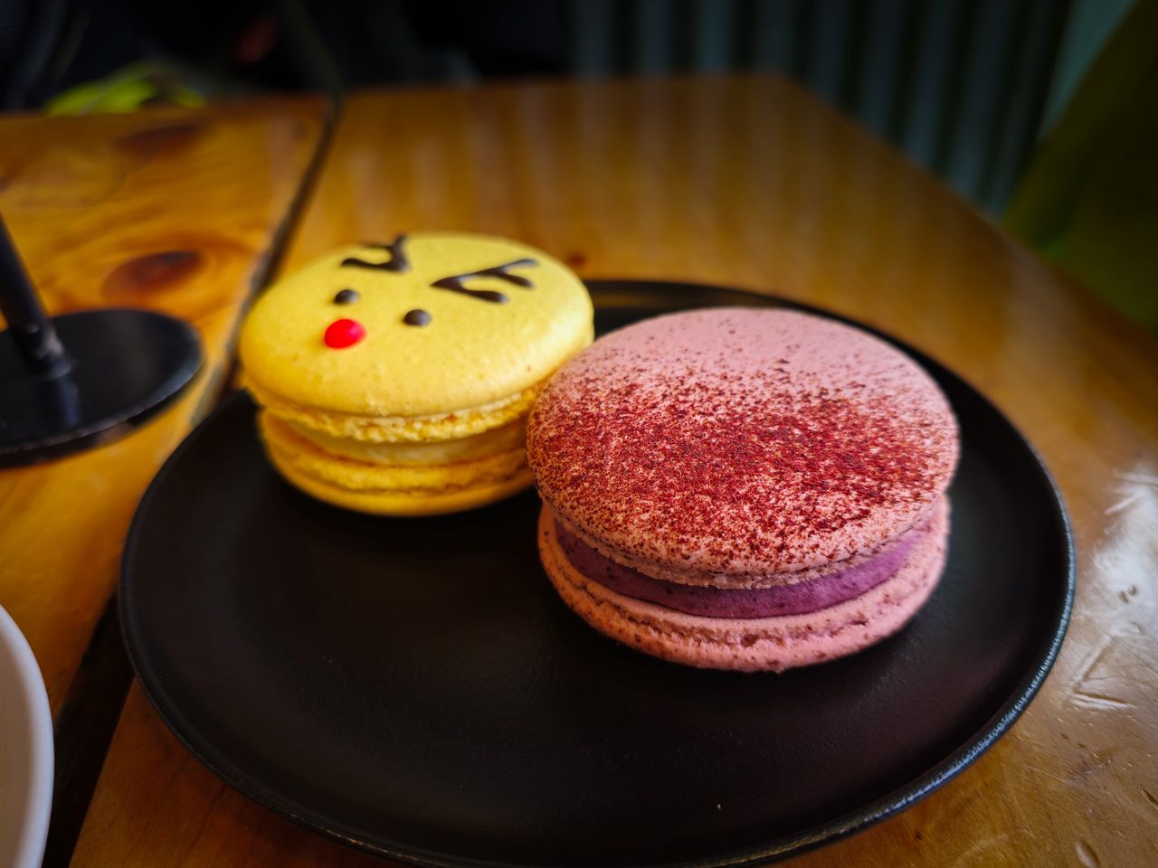 Two large macarons on a black plate on a wooden café table, one yellow with a cartoon reindeer face and one pink dusted with red powder