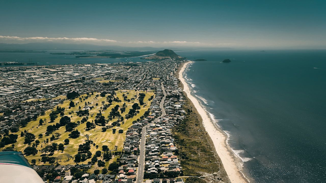 Aerial view along a long beach and seaside suburb to Mauao, a volcanic cone at a harbour entrance; aircraft wing at left