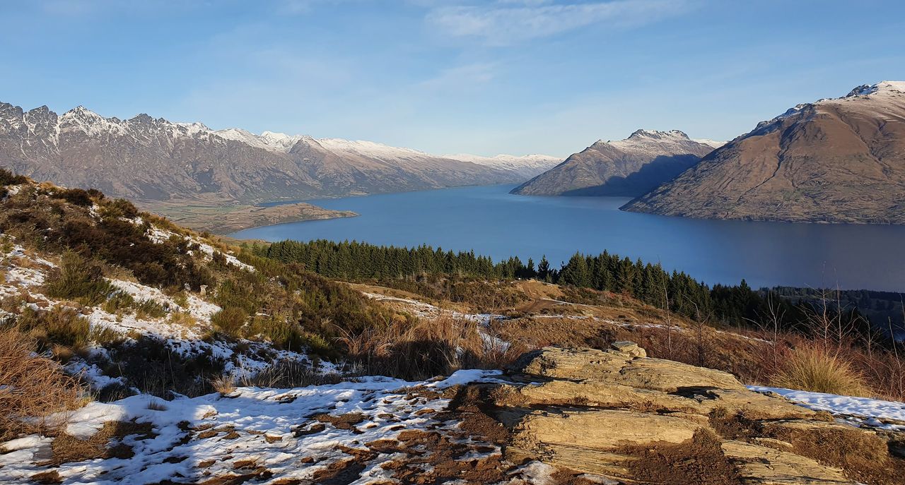 Lake Wakatipu from a snowy hillside above pine forest, the snow-dusted Remarkables on the left and steep peaks across the water