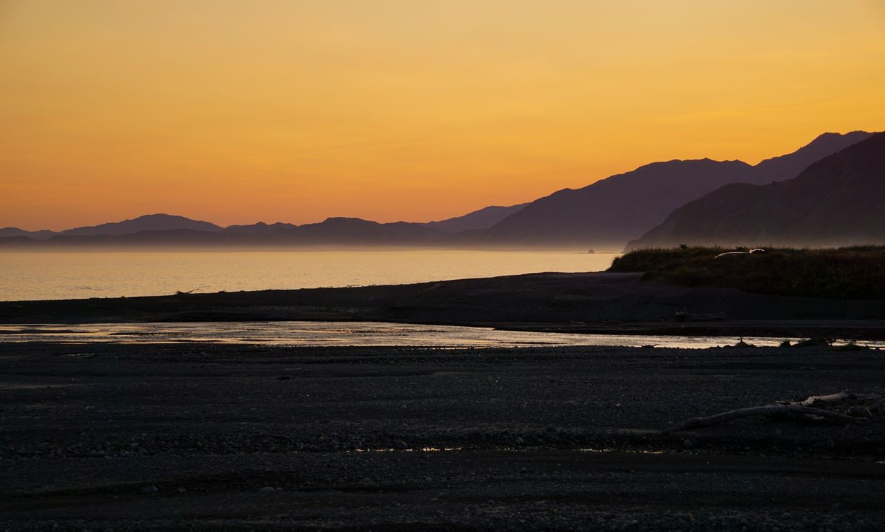 Orange sunset over calm sea and layered coastal ridges, with a shallow river channel crossing a dark stony beach
