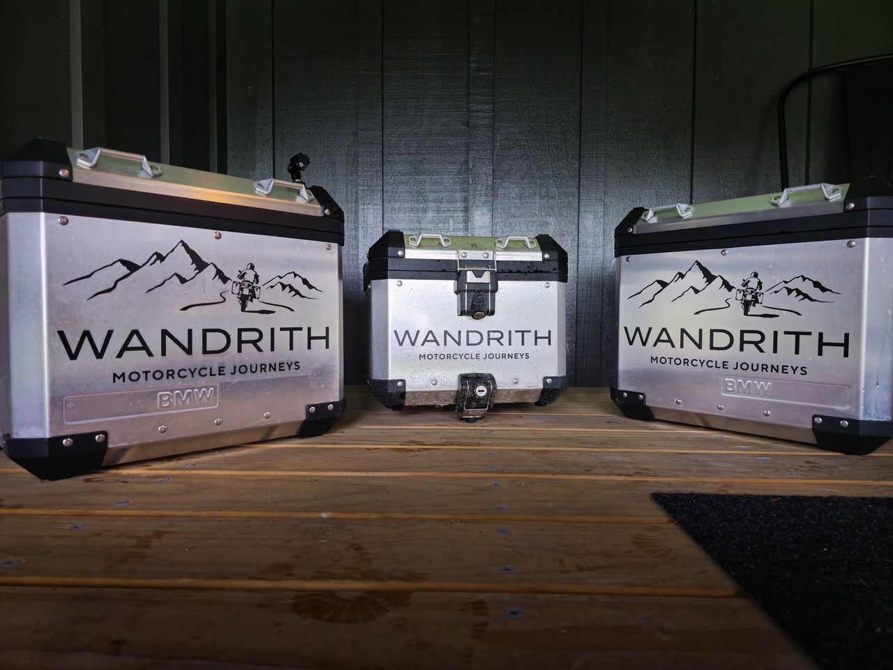 Two silver BMW panniers and a matching top box, each with the Wandrith Motorcycle Journeys logo, on a timber deck